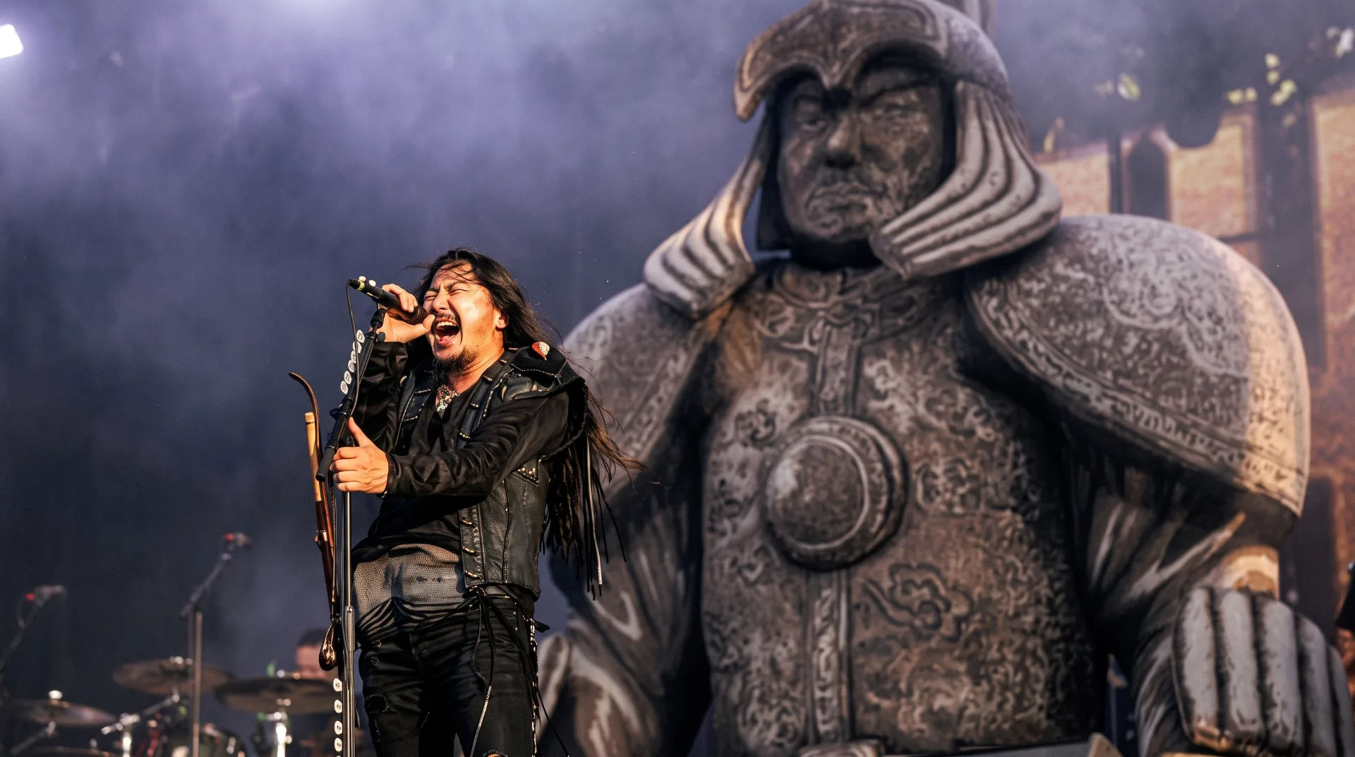 A singer passionately performing on stage with a large warrior statue in the background.
