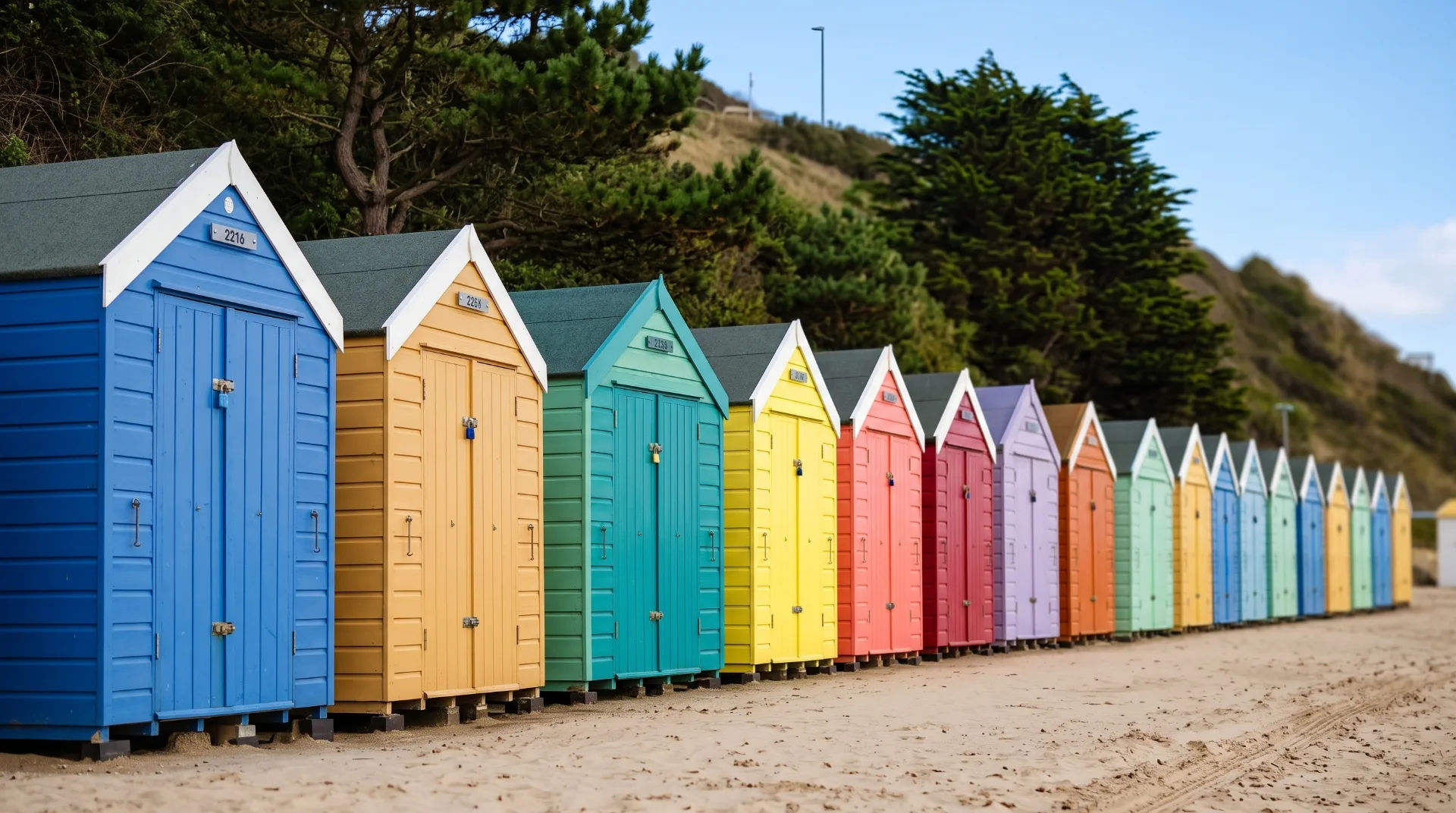 Row of colourful beach huts along sandy shore with greenery in background