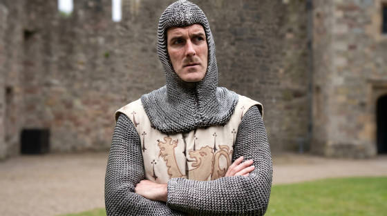 A man in chainmail and a tunic stands with arms crossed
