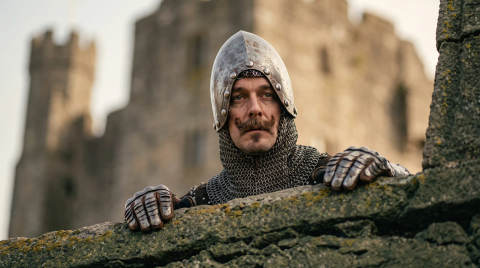 A knight in chainmail and helmet peering over a stone wall
