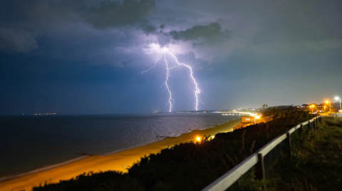 Lightning strikes over the sea with a beach and lights in the foreground
