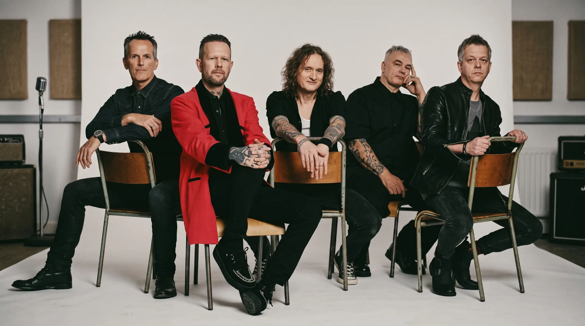 Five band members seated on wooden chairs, dressed in black and red attire, with a neutral background.