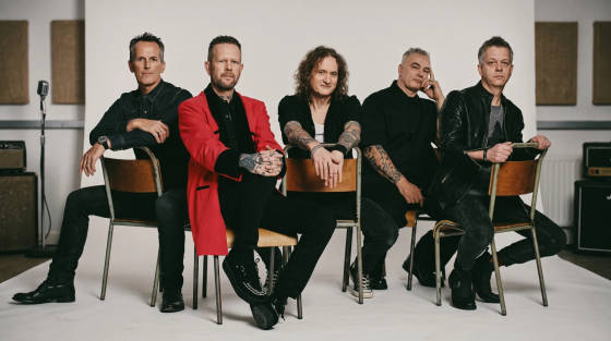 Six band members seated on chairs in a studio