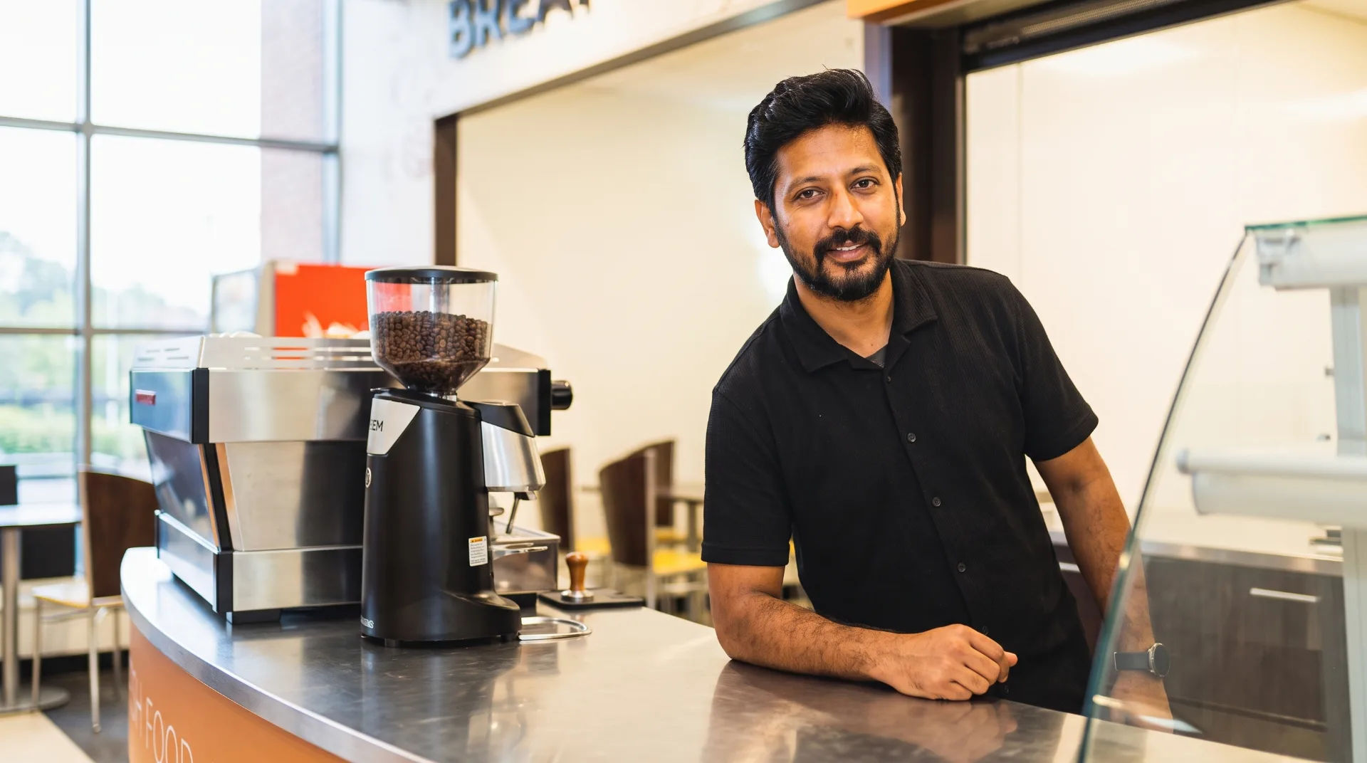 A man in a black shirt leans on a counter with coffee equipment in a bright restaurant setting.