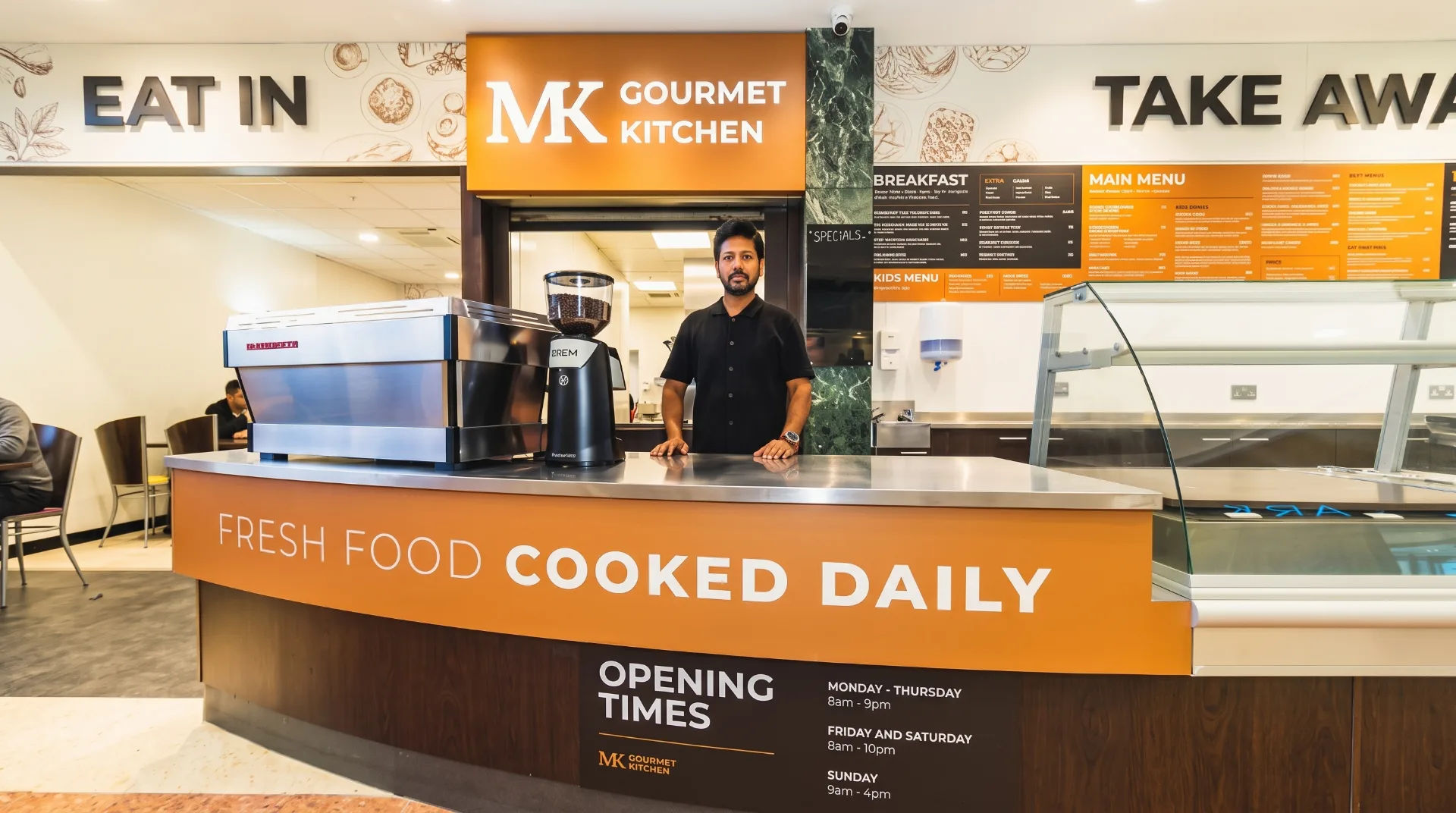 A man stands behind the counter of MK Gourmet Kitchen, showcasing the restaurant's menu and sign.