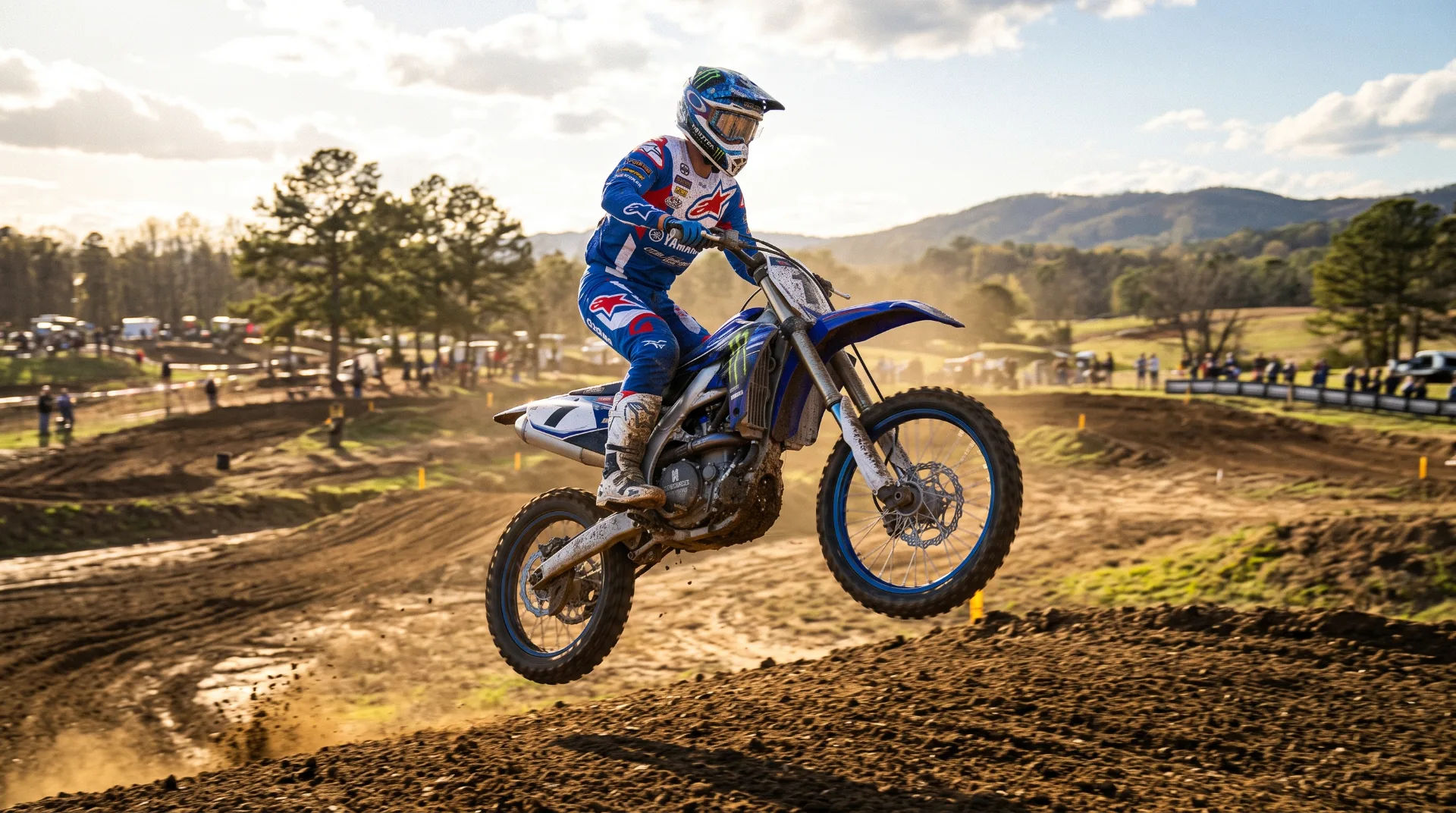 A motocross rider in blue gear jumps over a dirt mound during a race, with trees and spectators in the background.