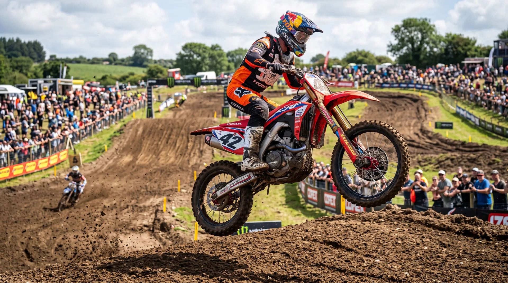 A motocross rider in orange and black gear jumps over a dirt mound, with spectators in the background.