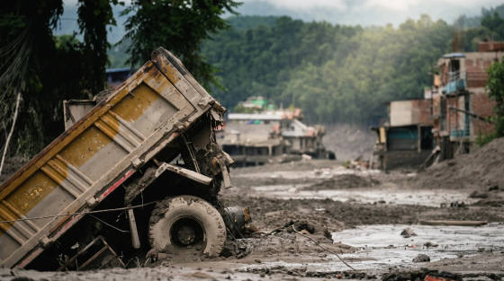 A tilted, muddy truck in a desolate area with buildings and boats in the background