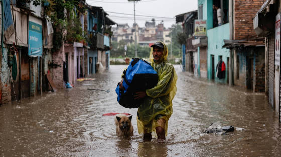 A man in a yellow raincoat carries a blue bag while walking through floodwaters with a dog