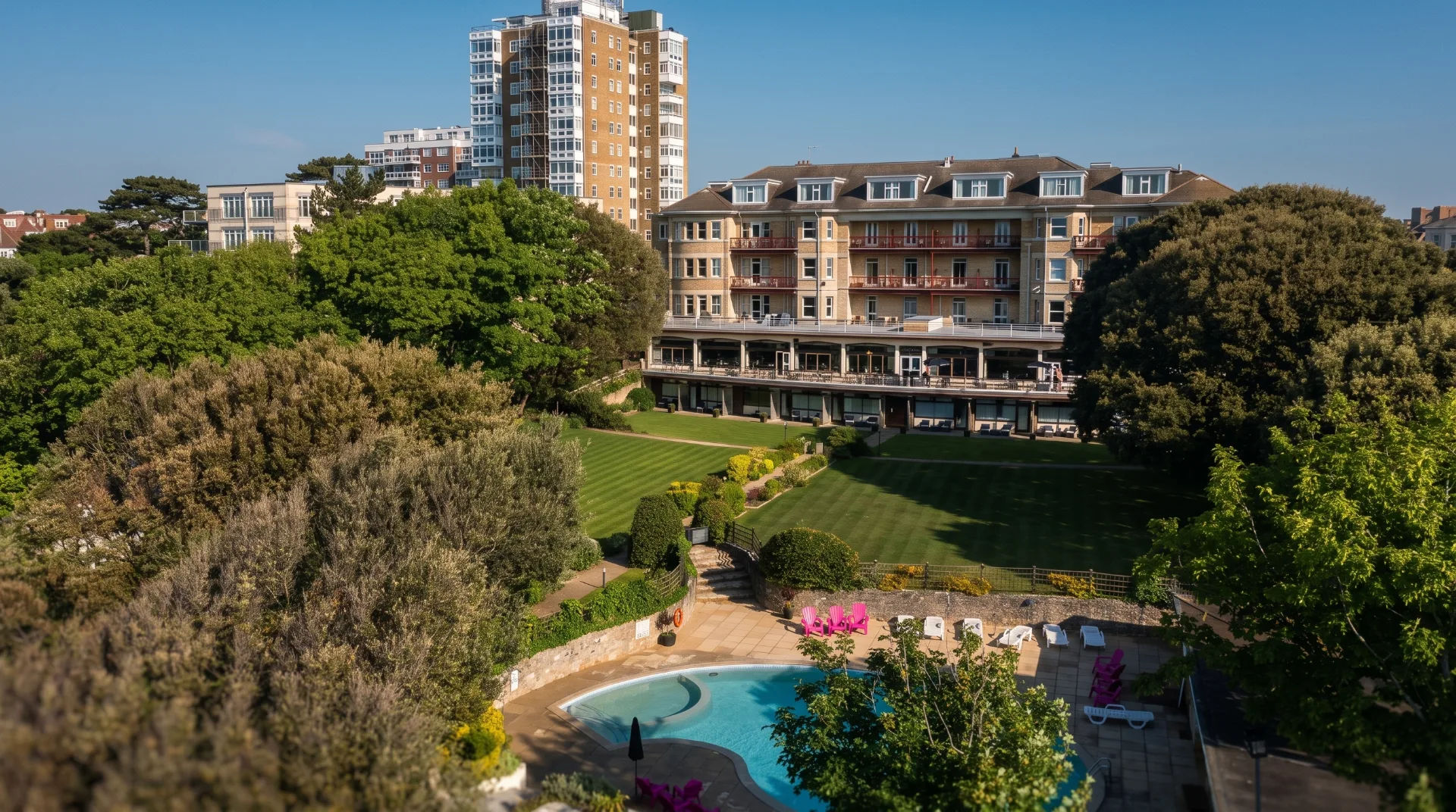 Lush green gardens with a swimming pool and a large building in the background, featuring balconies and modern architecture.