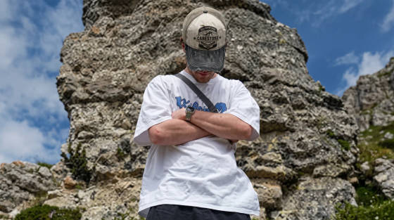 A young man in a white t-shirt and cap stands with arms crossed near rocks