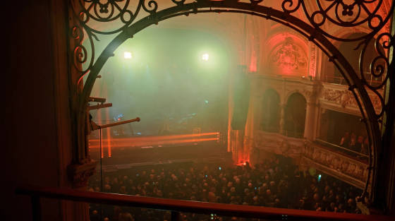 Concert scene viewed through an ornate archway with crowd and stage lights