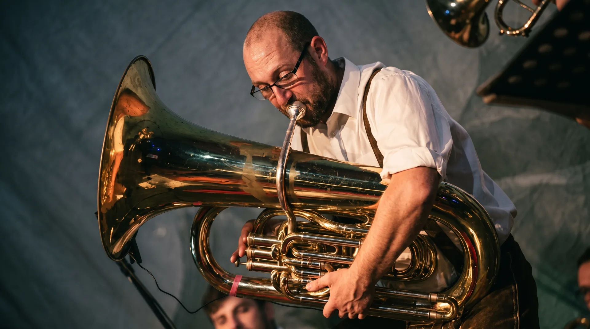Musician playing a large tuba with a focused expression, wearing glasses and a white shirt, with a blurred band member in the background.