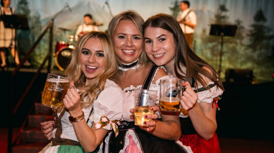 Three women in traditional outfits holding drinks and smiling