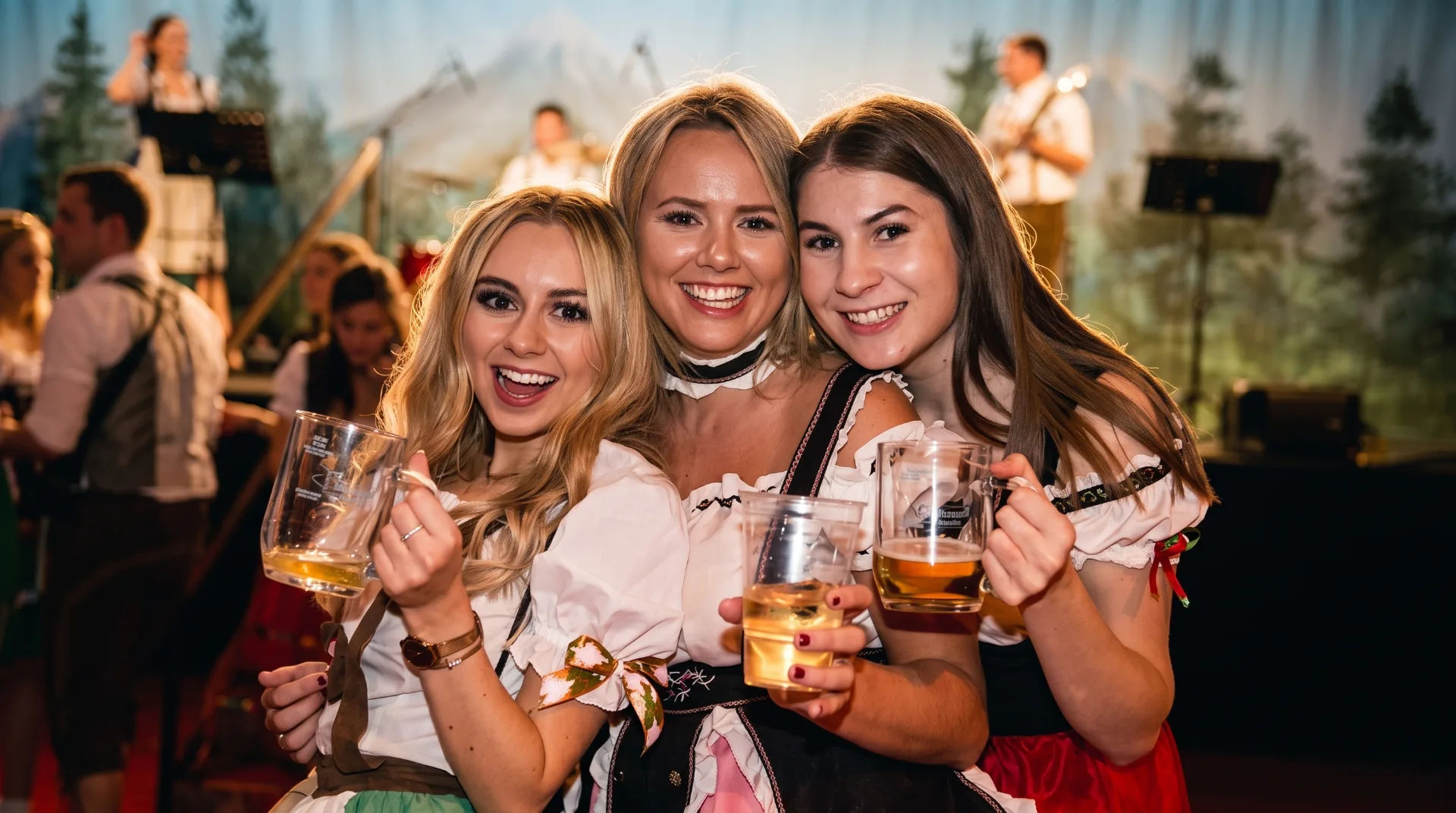 Three women in traditional Bavarian dresses holding beer mugs, smiling at the camera with a band in the background.