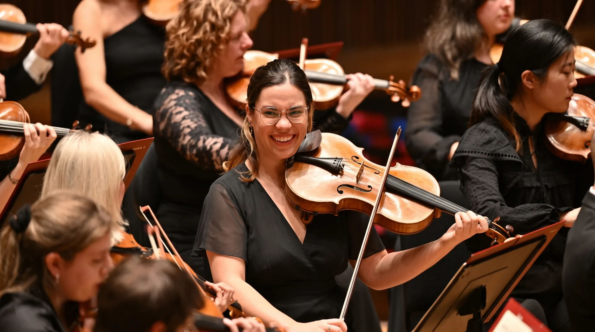 Violinist smiling while playing in an orchestra, with fellow musicians in the background