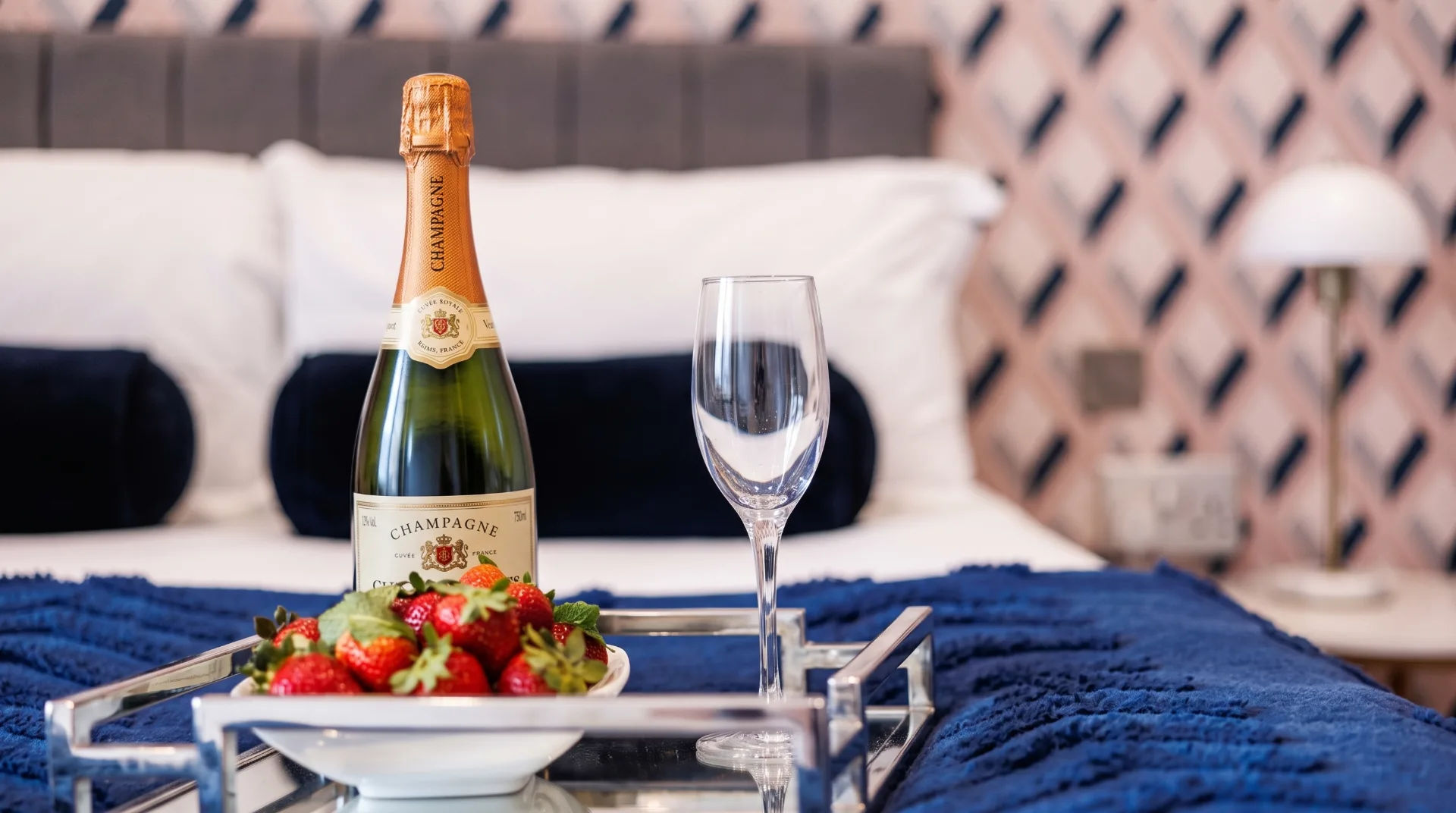 A bottle of champagne and a glass on a tray with strawberries, set on a blue blanket in a stylish bedroom.