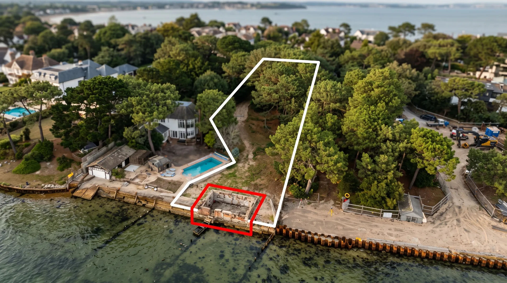 Aerial view of a coastal area with a pool, trees, and a marked structure near the water.