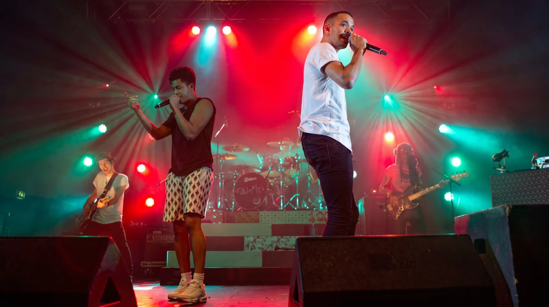 Two male performers on stage with microphones, vibrant lights in the background, a guitarist visible behind them