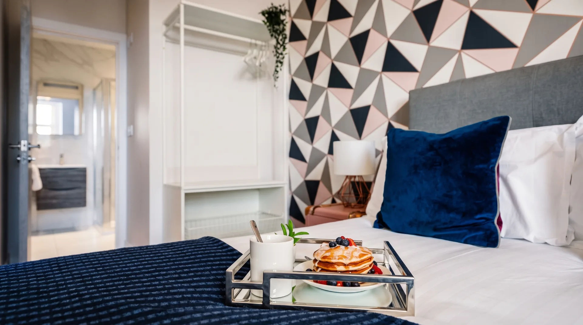 King-size bed with blue cushions, breakfast tray featuring pancakes and berries, geometric patterned wall
