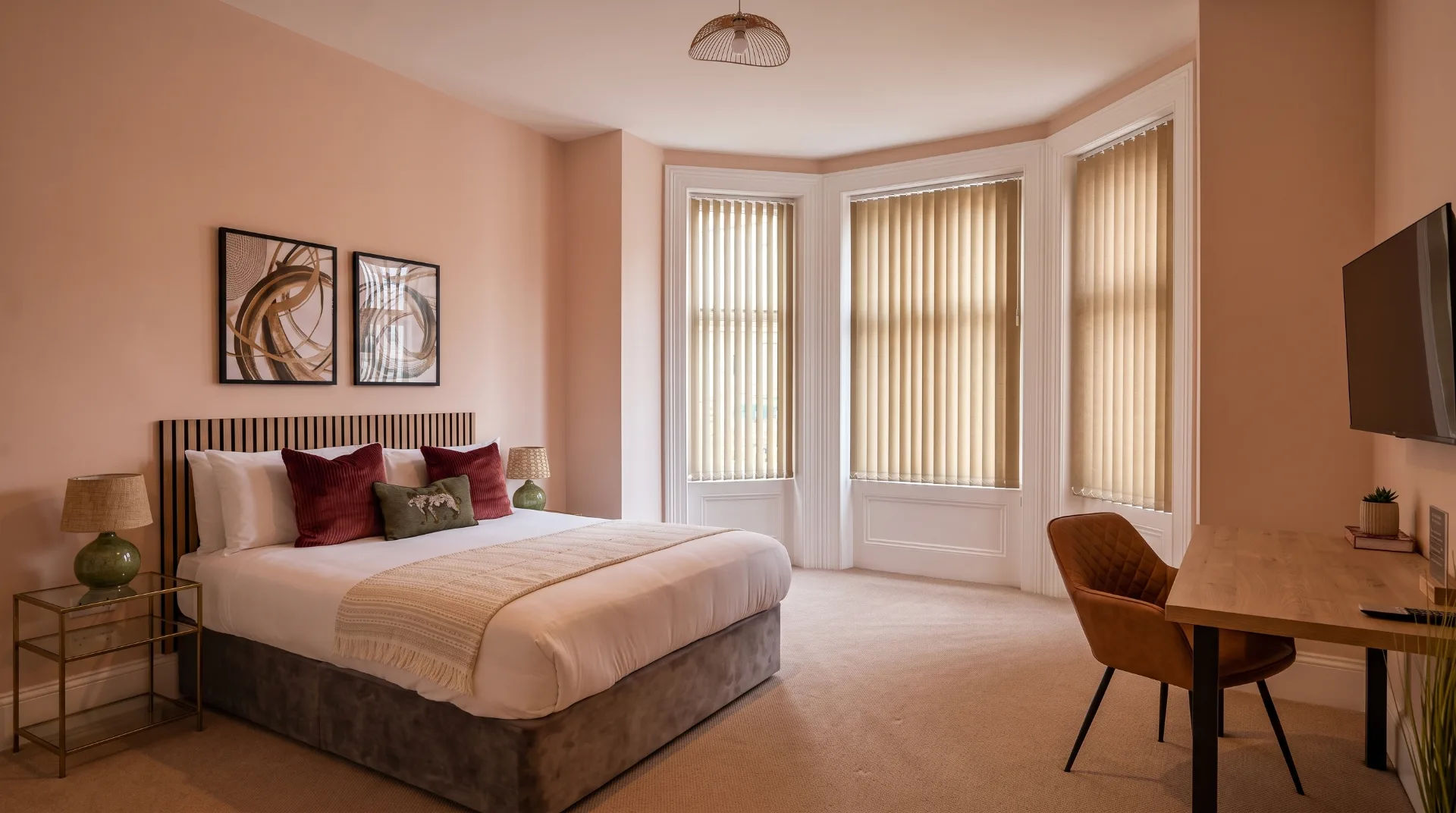 Stylish bedroom with a double bed, decorative cushions, and a desk in a light-coloured room with large windows.