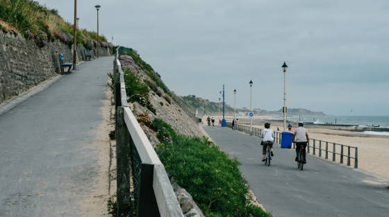 Two cyclists riding along a coastal path with benches and flowers