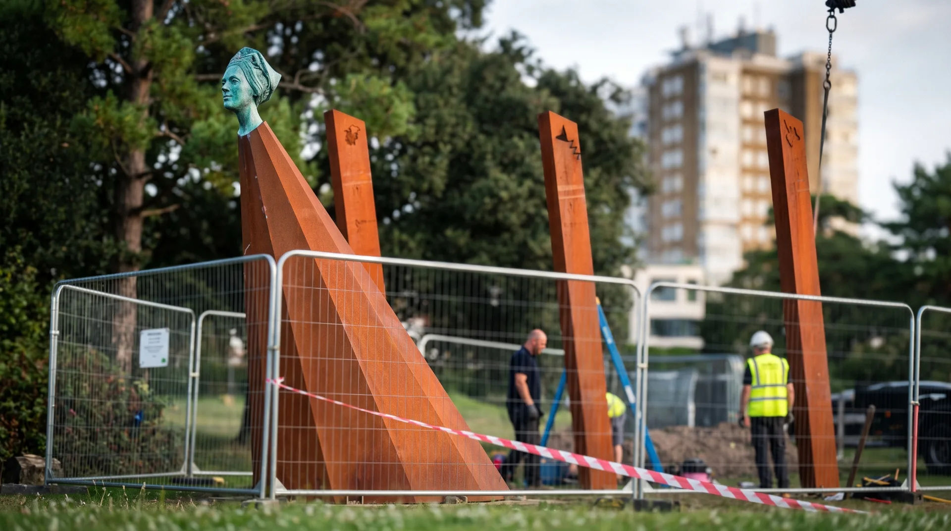 A contemporary sculpture featuring a green head atop angular brown structures, surrounded by construction barriers and workers