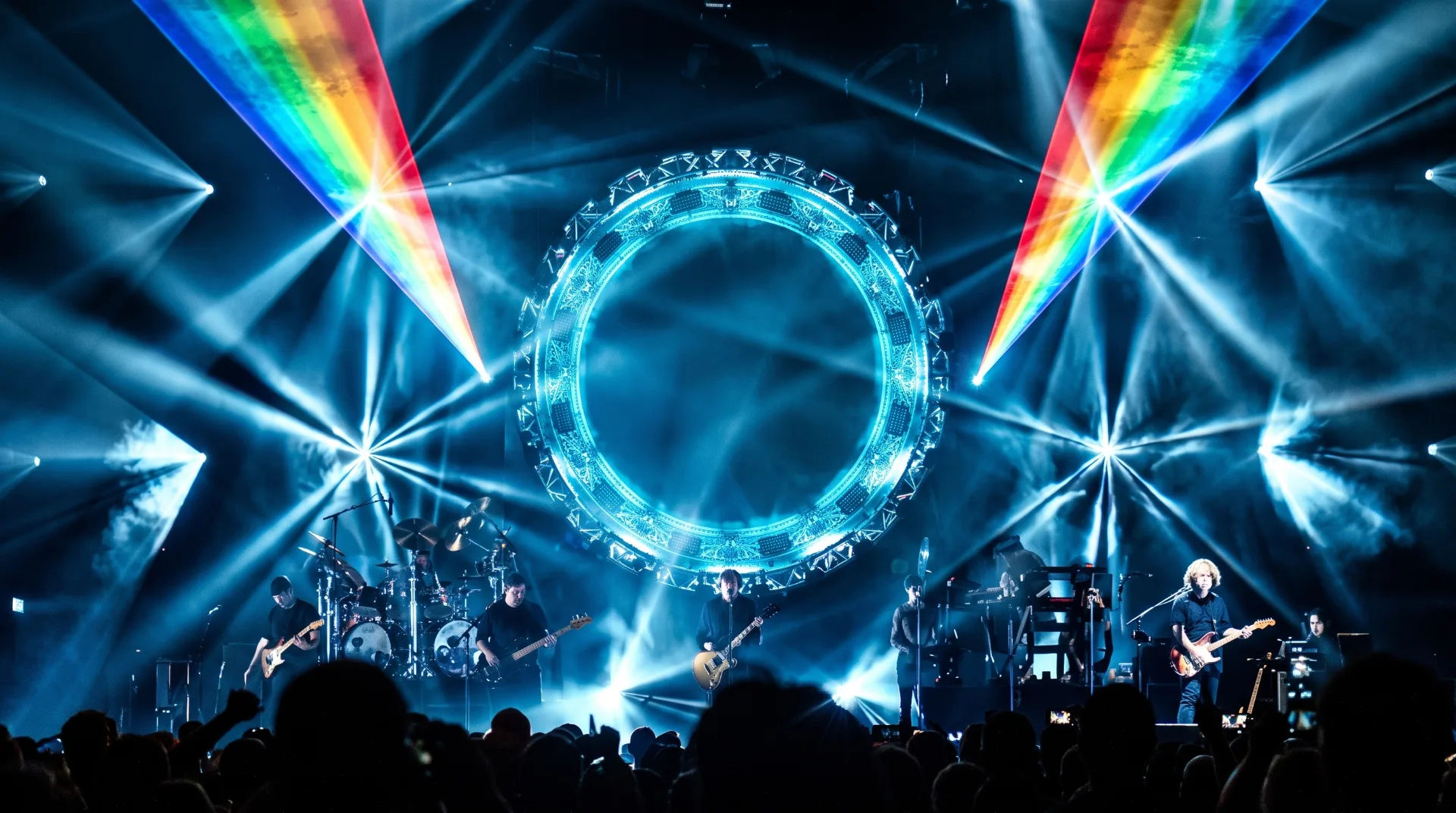 Bright stage lights and laser effects illuminate a band performing with an audience in silhouette.
