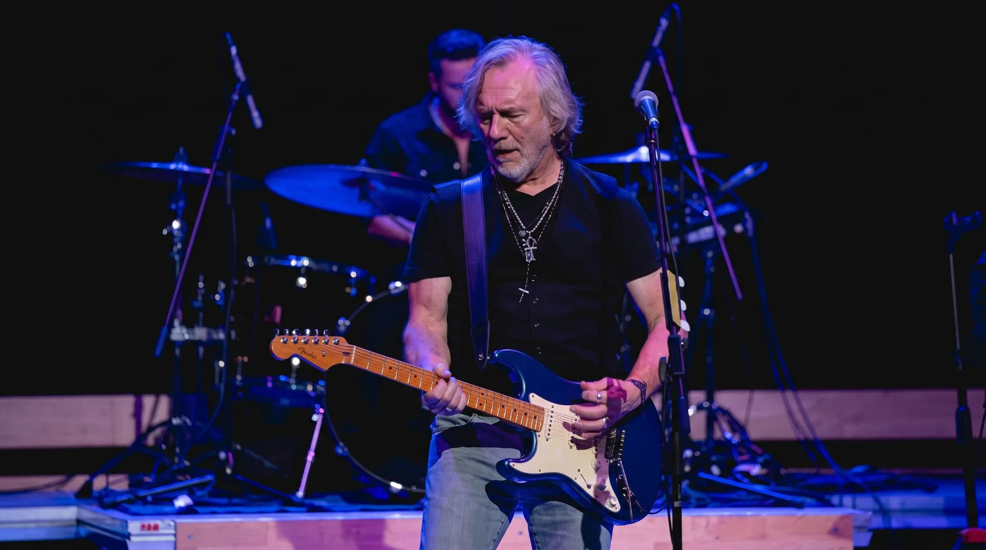 Musician performing with an electric guitar on stage, drums visible in the background