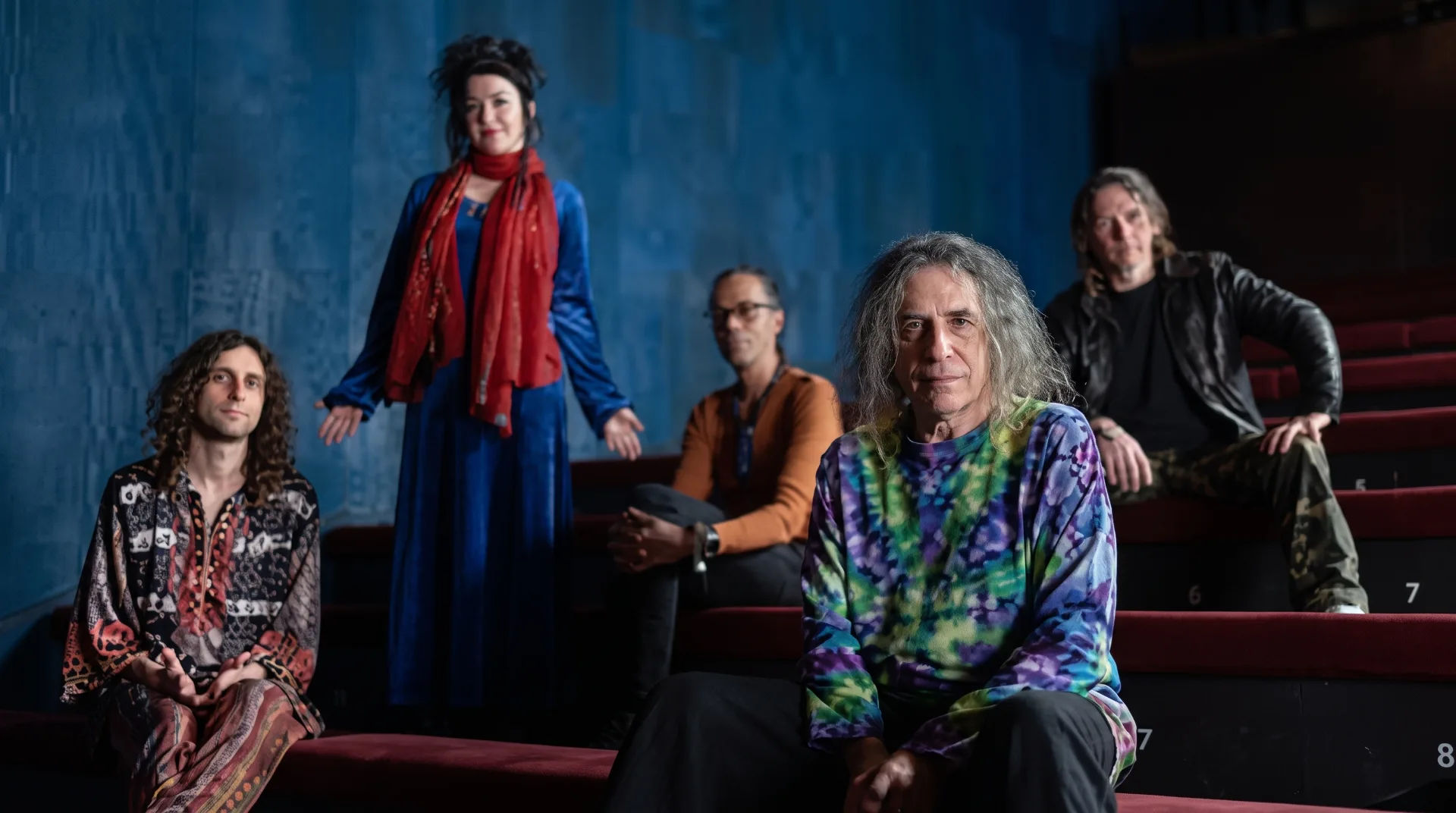 Five musicians posed on red seating against a blue wall, showcasing vibrant outfits and unique styles.