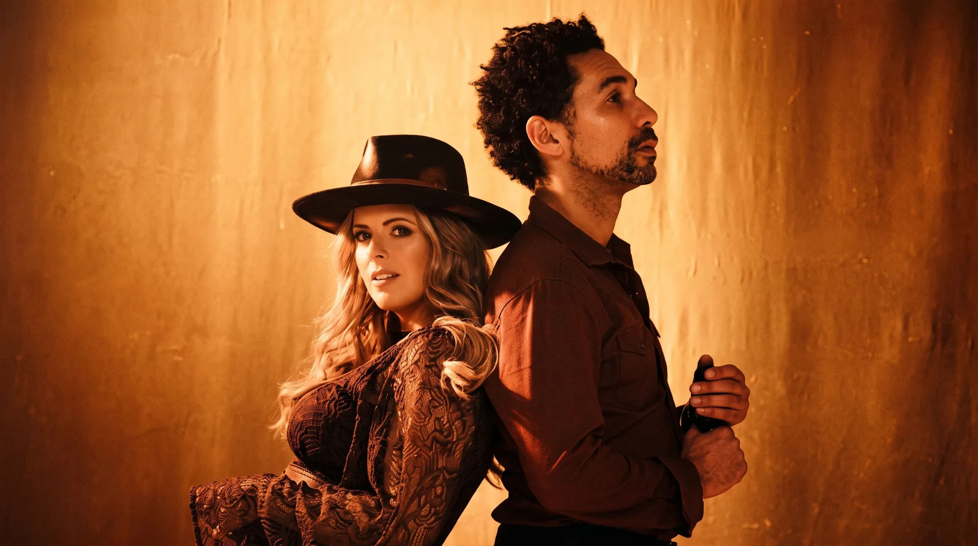 A man and woman pose back to back against a warm, golden backdrop, both dressed stylishly.