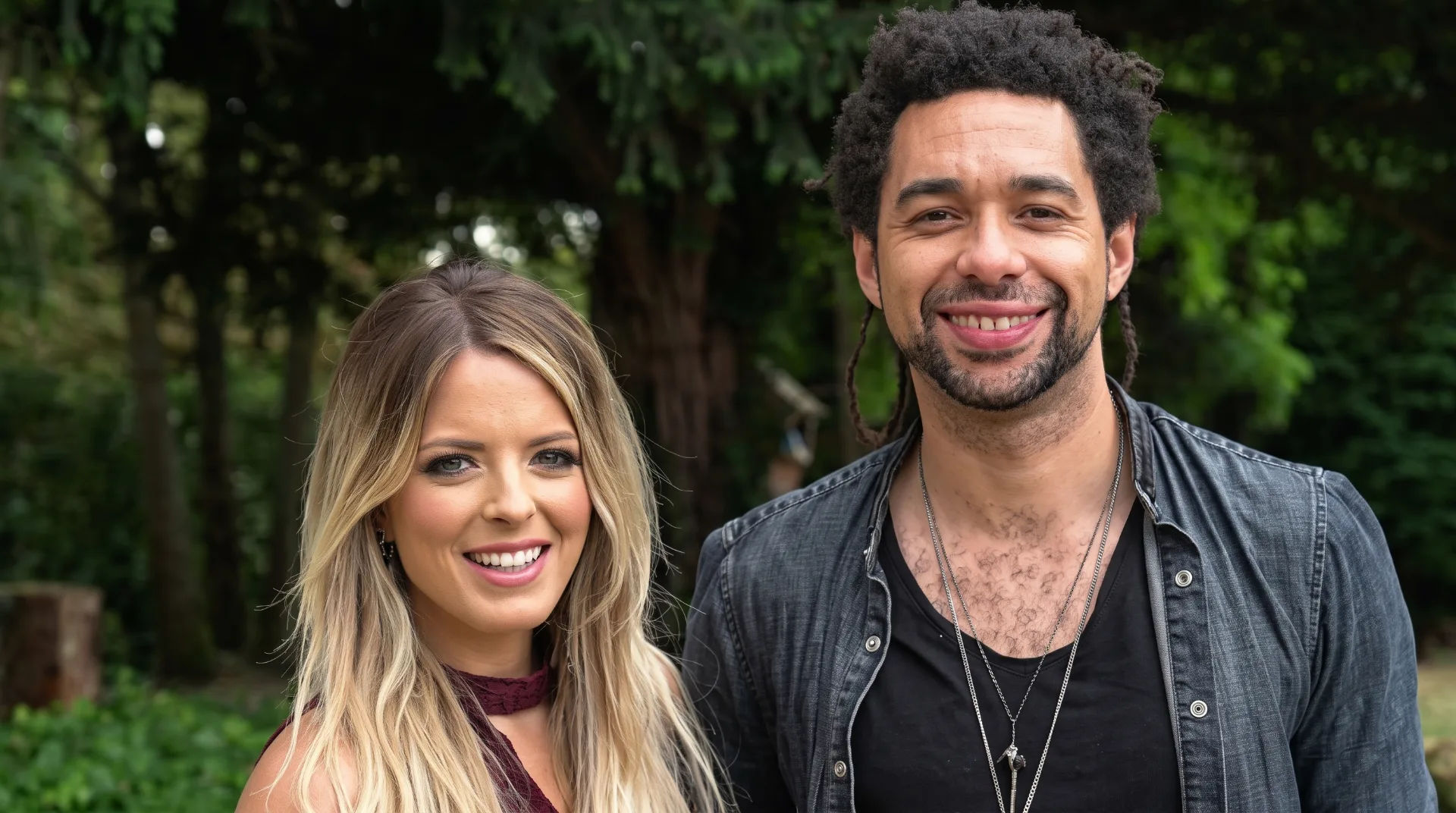 Two smiling musicians stand outdoors, surrounded by greenery, with one wearing a black shirt and the other in a maroon top.
