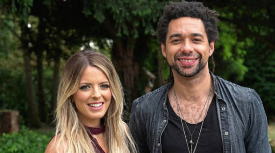 Two smiling individuals standing outdoors with greenery behind them