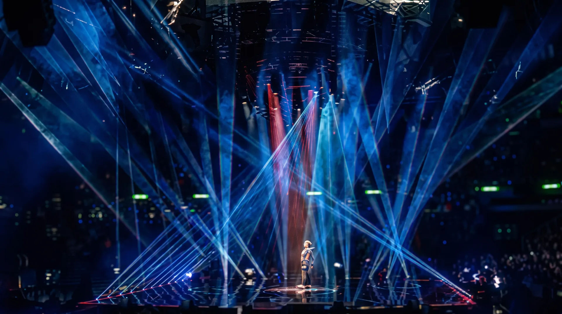 A performer stands on stage surrounded by vibrant blue and red laser lights at a concert.