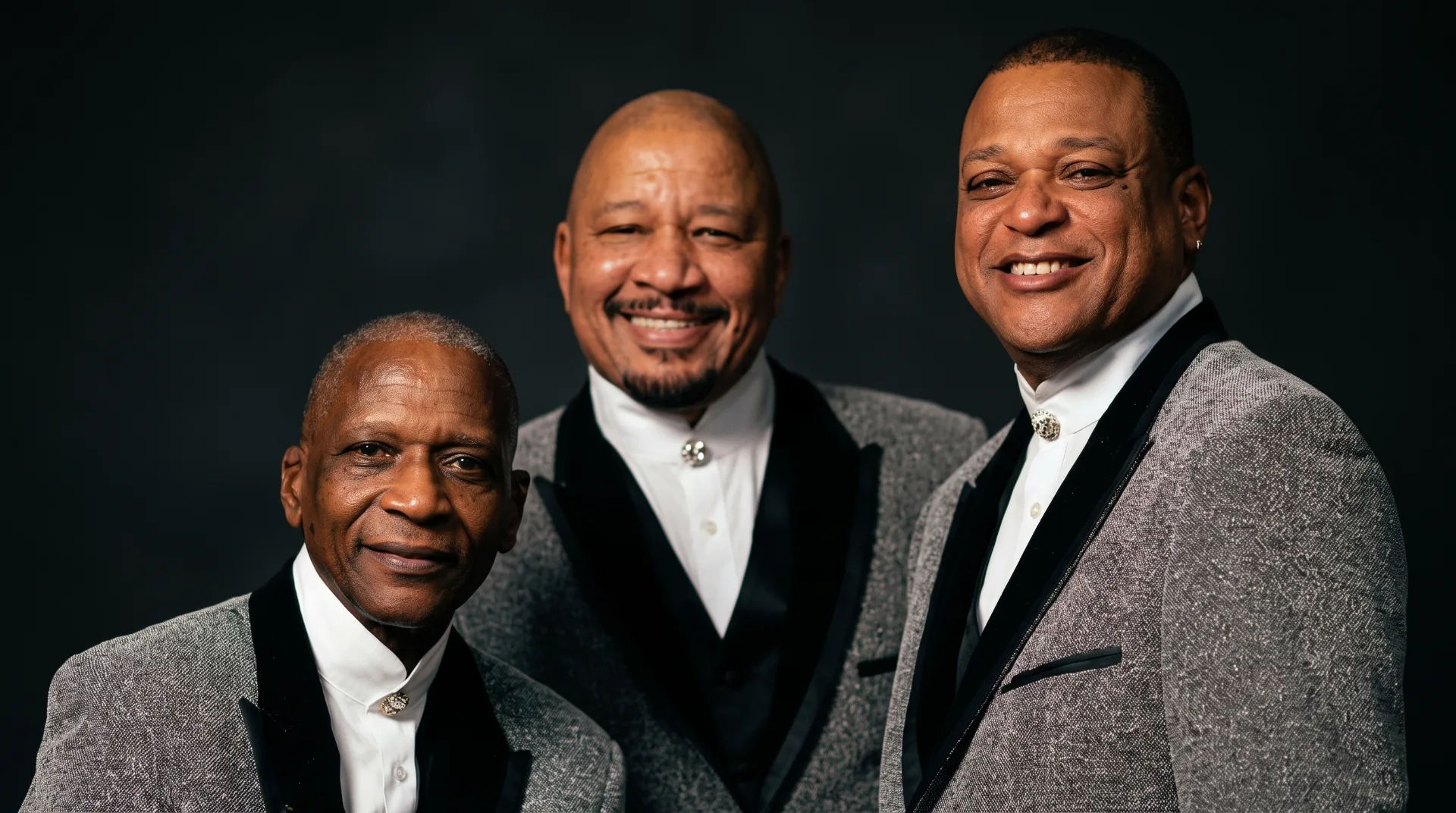 Three male singers in stylish grey suits with black accents, smiling against a dark background