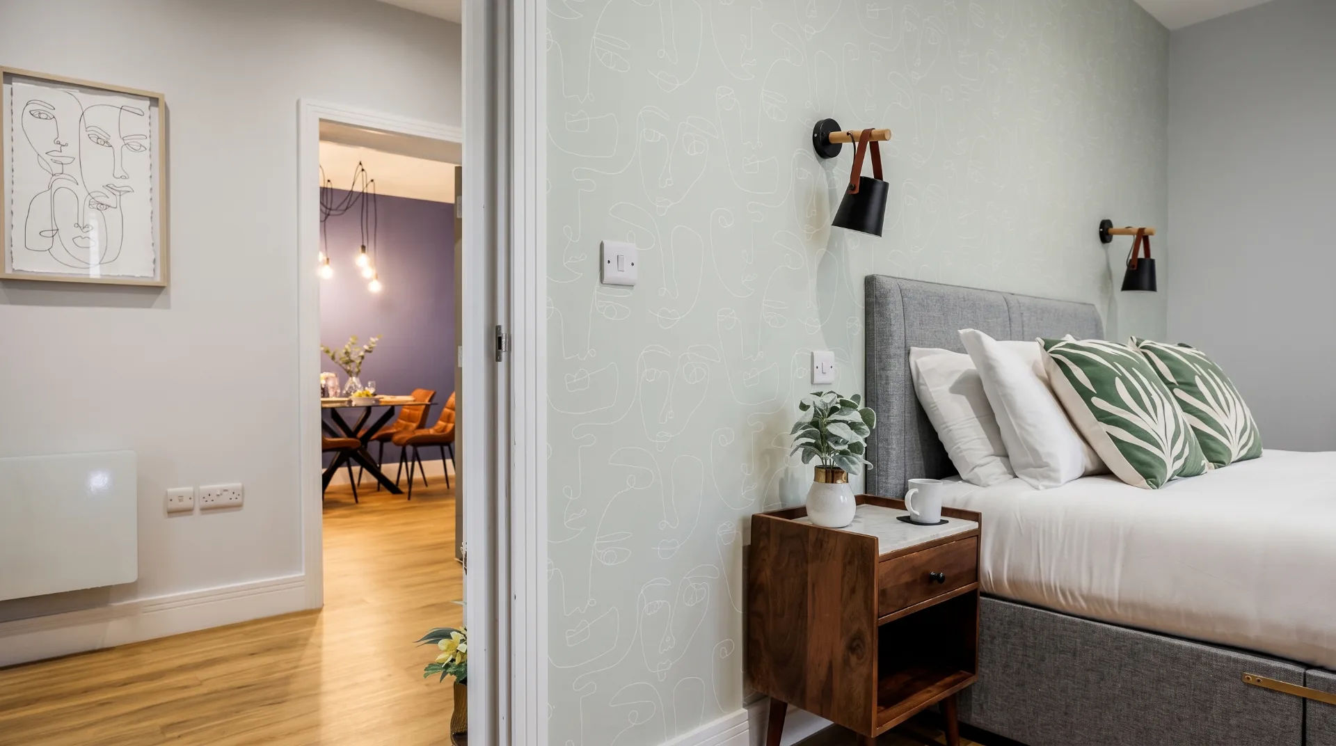 Stylish bedroom with grey bed, leafy cushions, and modern decor, leading to a dining area.