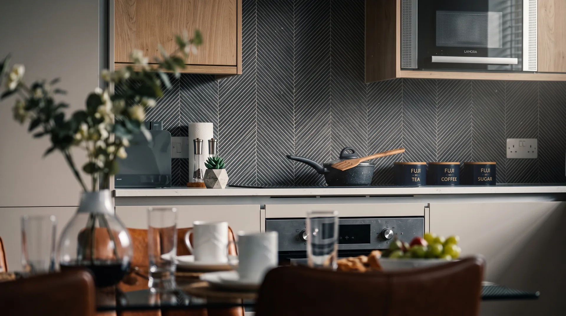 Modern kitchen with wooden cabinets, herringbone backsplash, and dining table set with cups and fruit