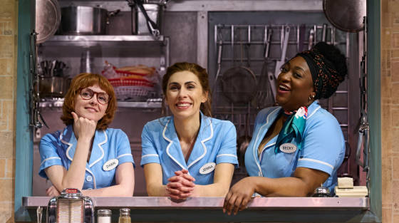 Three women in blue diner uniforms smiling in a kitchen