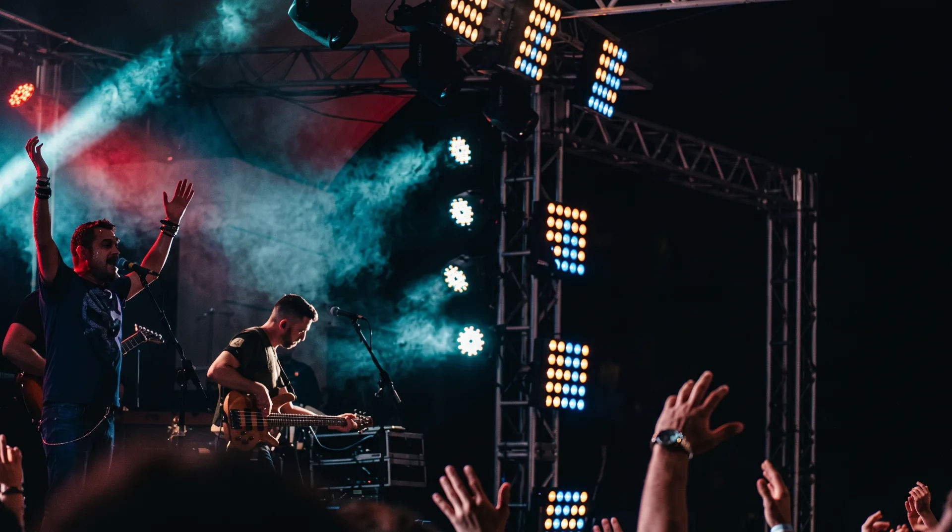 A lively performance with a singer gesturing and a bassist playing, surrounded by vibrant stage lights and smoke.