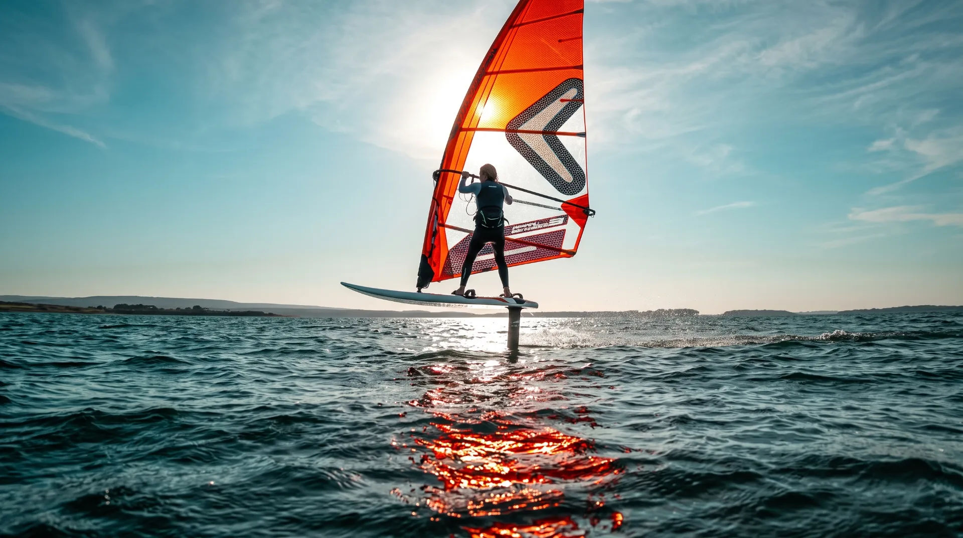 A windsurfer on a board with a bright orange sail gliding over shimmering water under a clear sky.