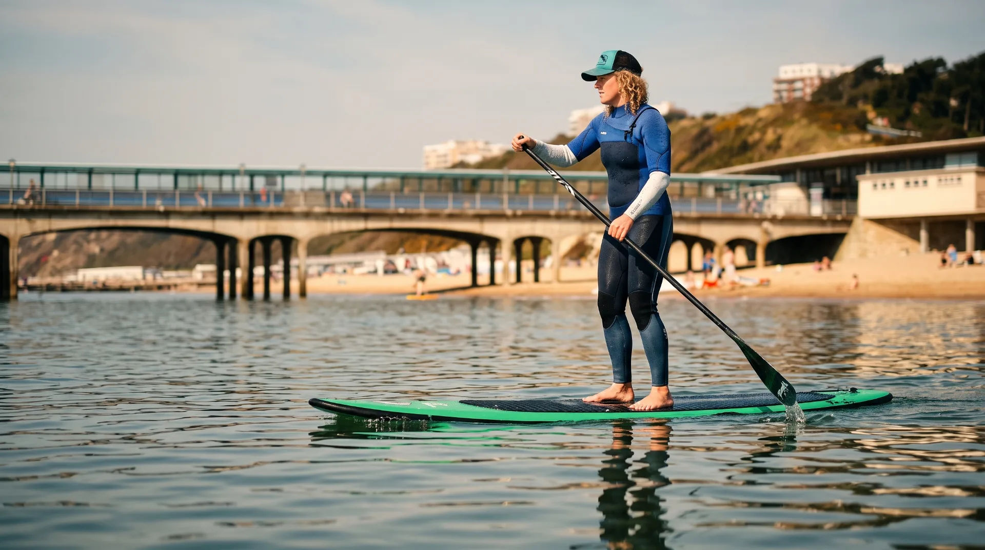 A woman paddleboarding on calm waters with a pier in the background and beachgoers on the shore.