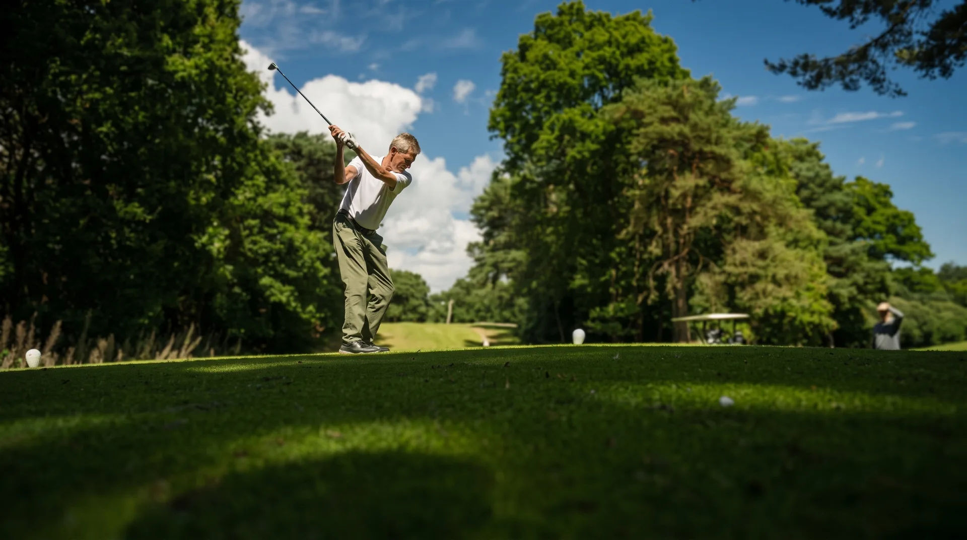 A golfer swings on a lush green course surrounded by trees under a blue sky with clouds.