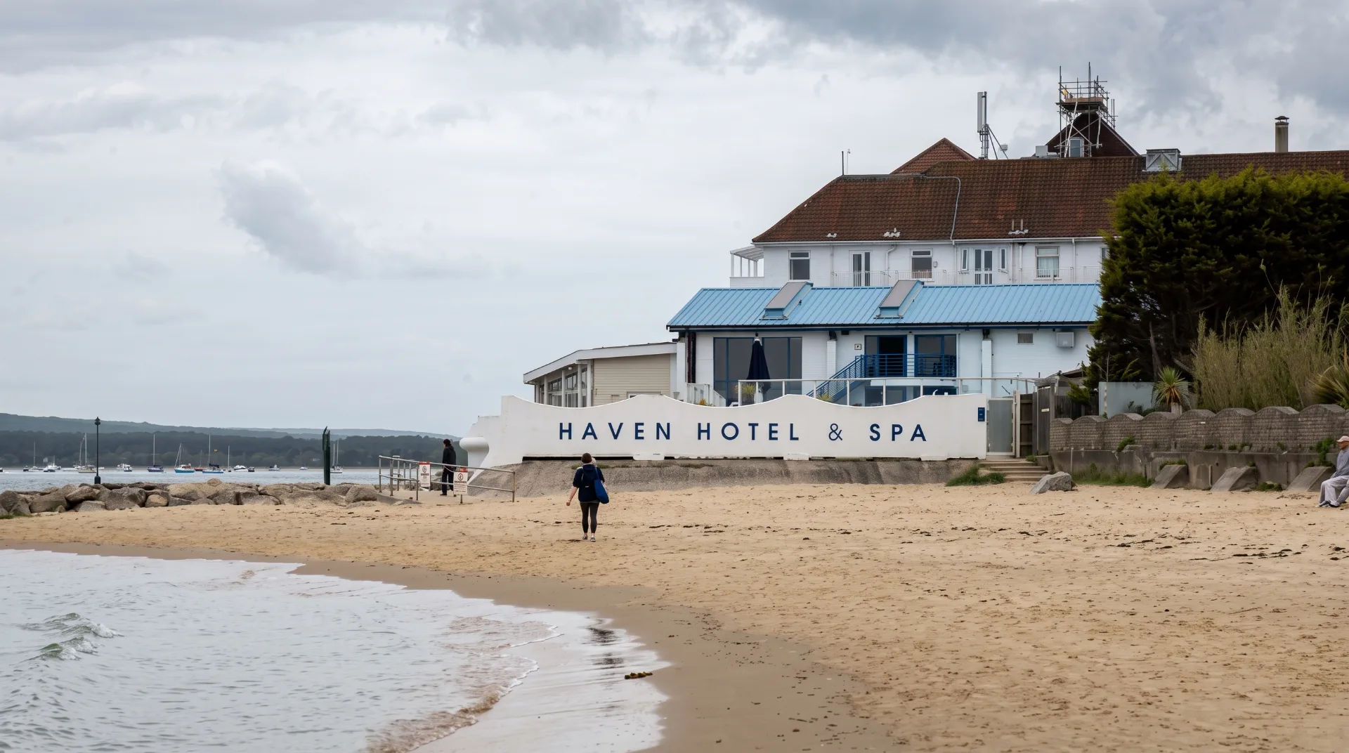 Haven Hotel & Spa on Sandbanks beach with a person walking along the shore and boats in the background