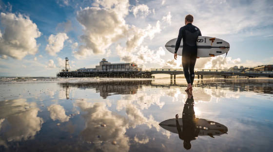 A surfer in a wetsuit carrying a surfboard by the water
