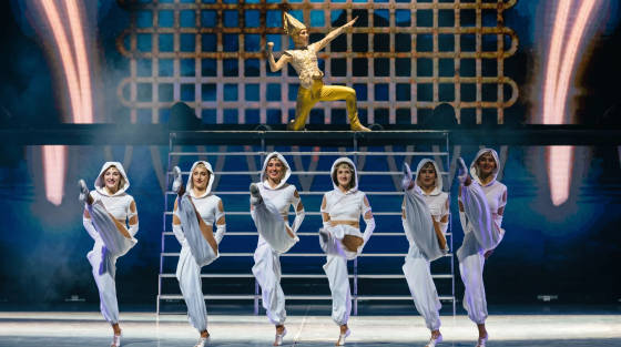 Dancers in white costumes performing on stage with a male performer in gold