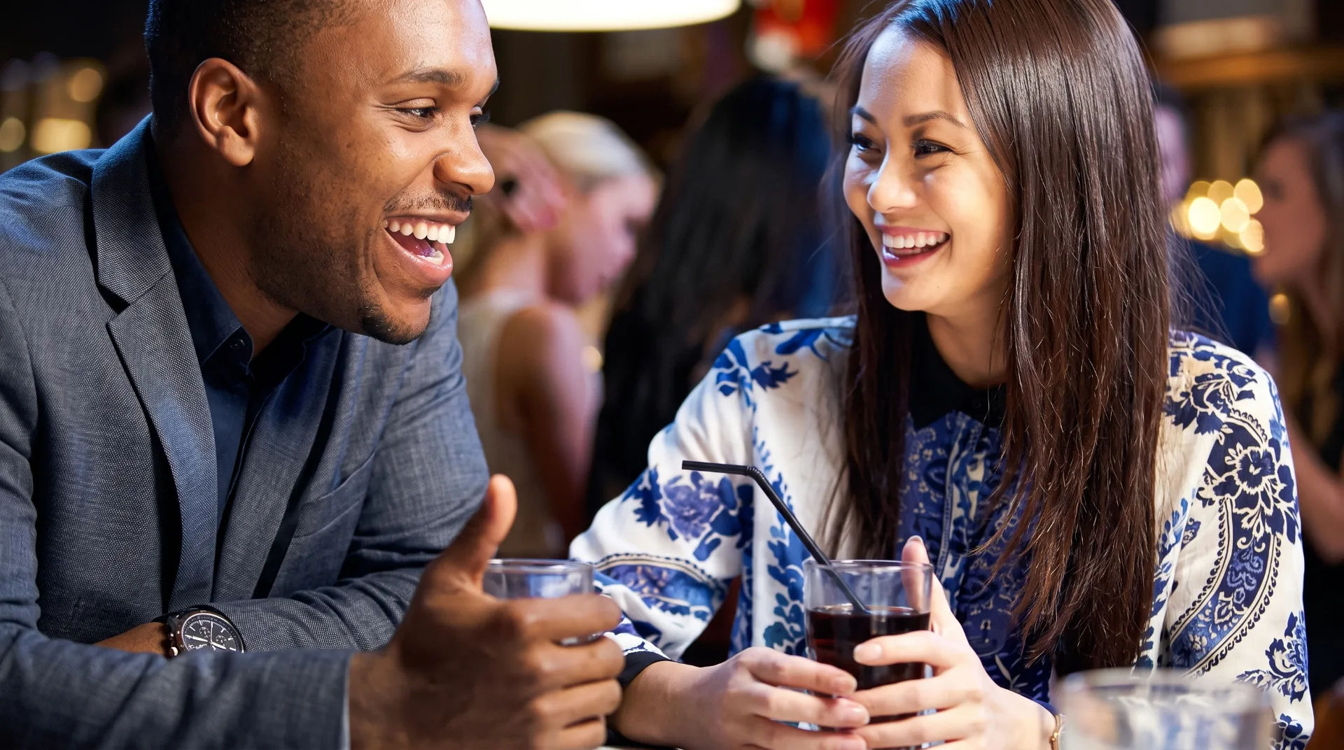 Two smiling individuals engaged in conversation at a bar, holding drinks, with a lively background