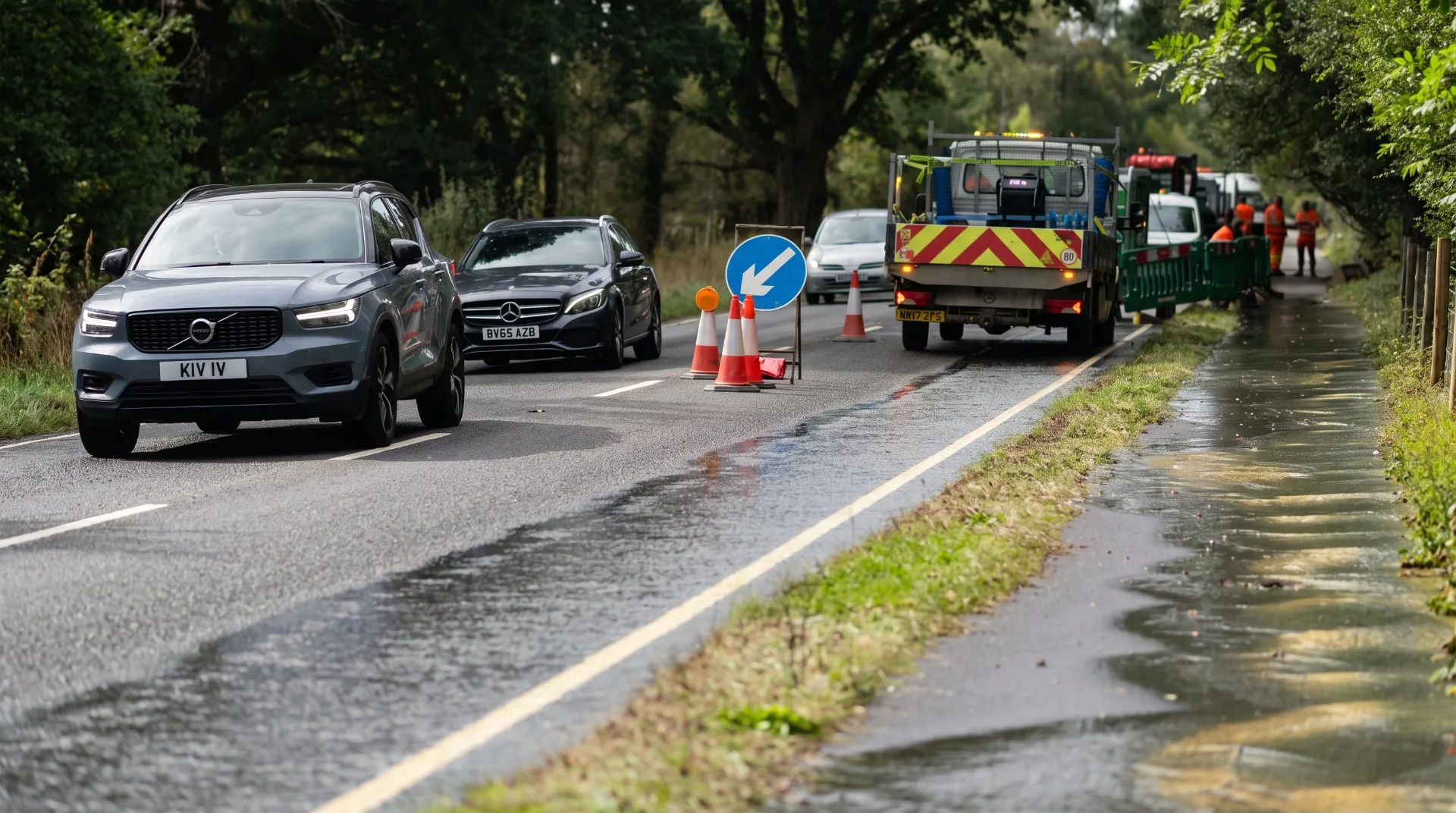 Traffic on a road with cones and a water leak on the surface, emergency vehicles present nearby