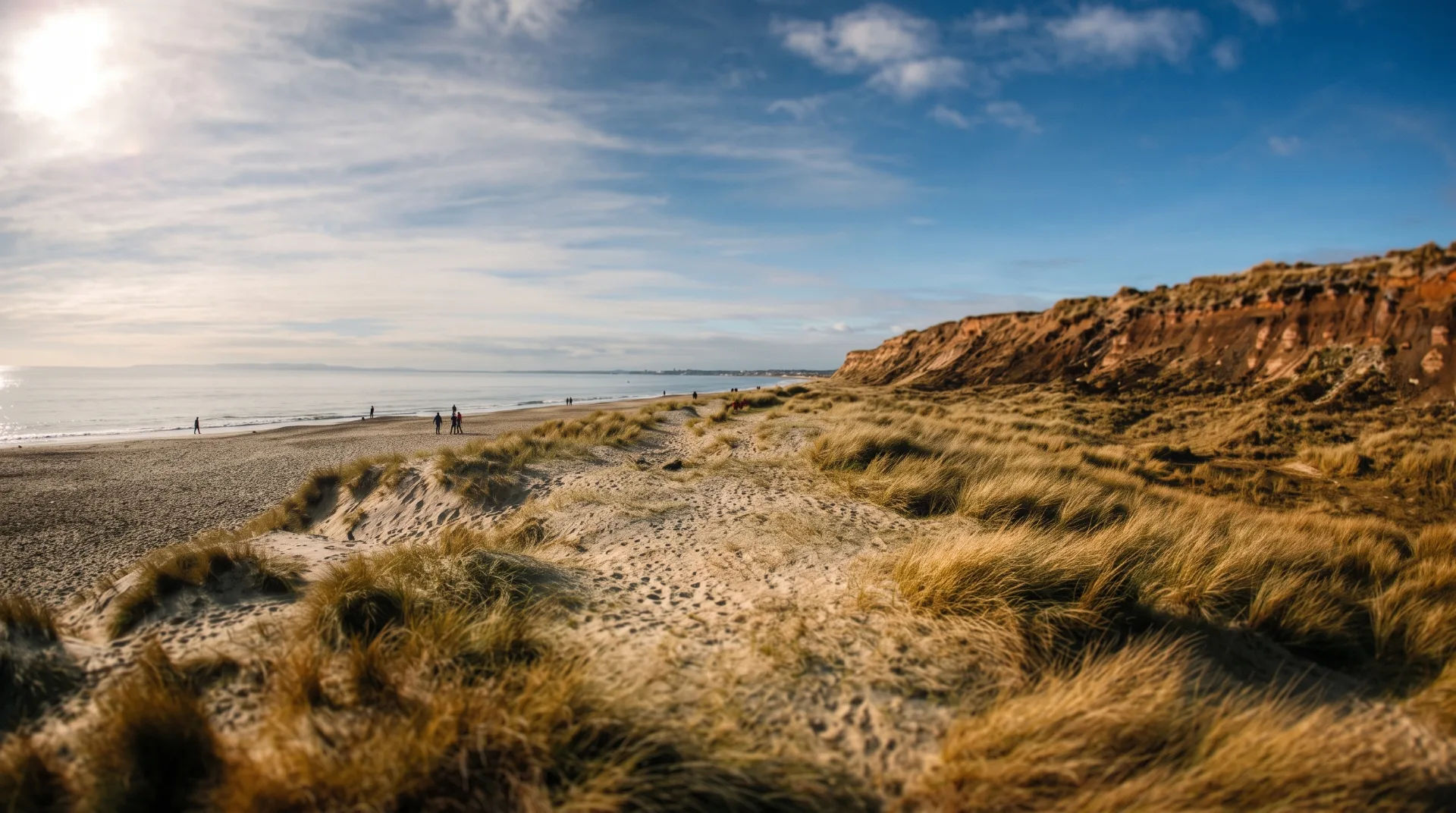 A sandy beach with tall grass dunes and cliffs in the background, under a blue sky with scattered clouds.