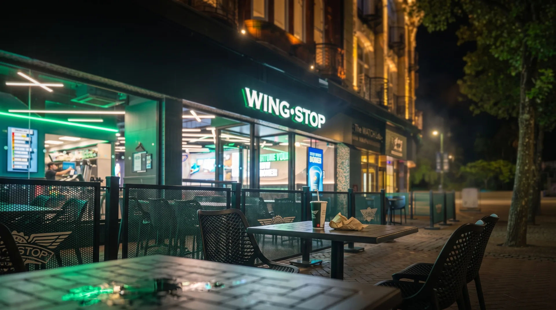 Exterior of Wingstop restaurant at night, featuring green neon lights and outdoor seating with tables and chairs.