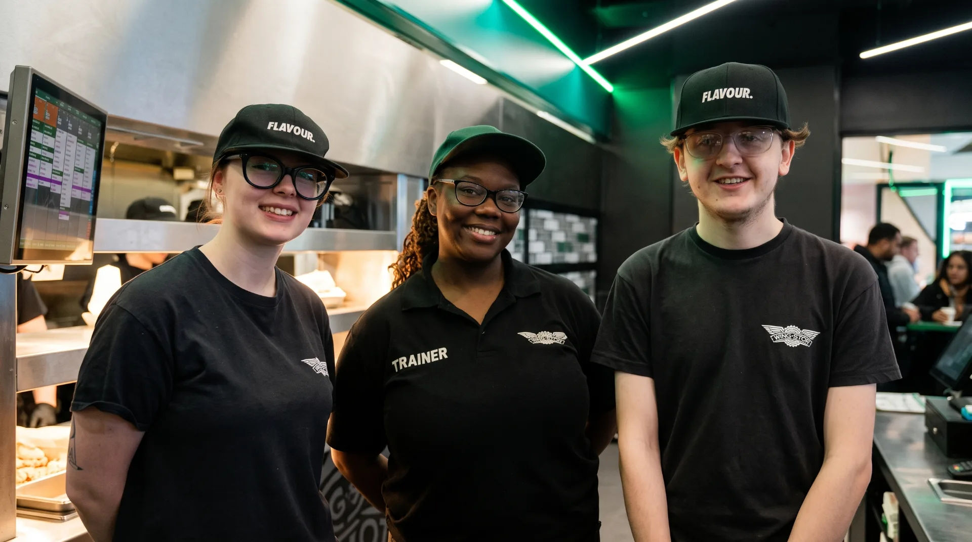Three staff members in black uniforms and caps stand in a restaurant, smiling at the camera.
